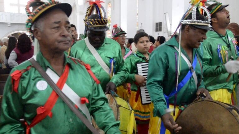 Membros de um grupo de Congados vestidos com trajes coloridos durante uma apresentação em um ambiente interno.